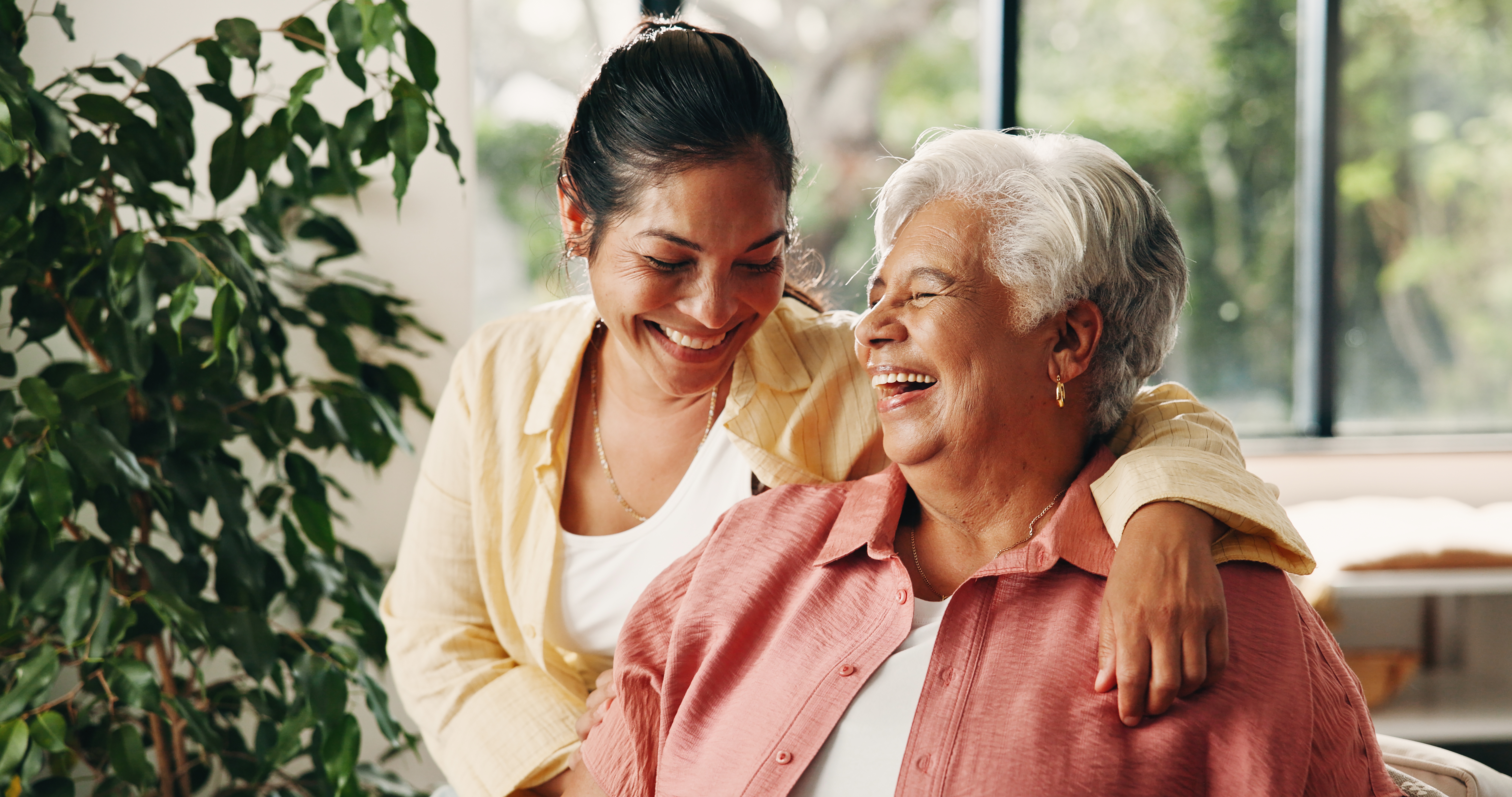 A younger woman with dark hair embraces an older woman with grey hair, both laughing joyfully indoors.