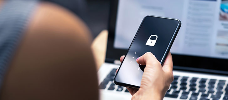 A hand holding a smartphone displaying a padlock icon and a numeric keypad, with a laptop in the blurred background.