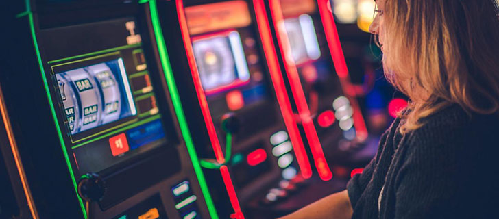 A woman with blonde hair plays a slot machine with 'BAR' symbols on its screen, surrounded by colorful red and green lights in a dark setting.