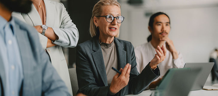 A blonde woman in glasses speaks and gestures during a meeting with colleagues, including a man listening intently with a laptop.