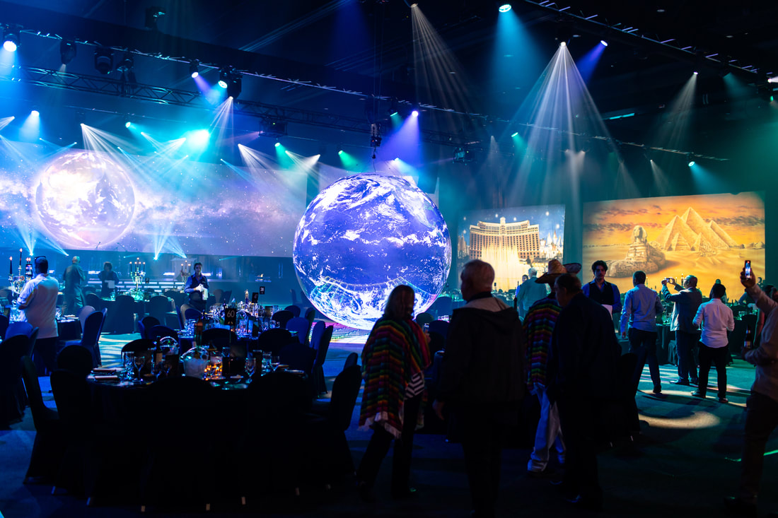 An event space with large spherical Earth projections, screens showing the Bellagio and pyramids, and people mingling under blue lights.