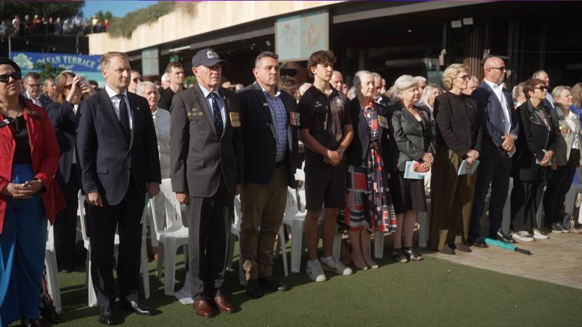 A group of people, including men in suits and some wearing medals, stand on green turf at an outdoor event.