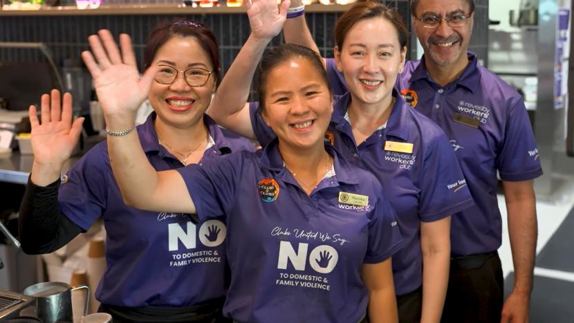 Four smiling people in purple polo shirts wave at the camera. Their shirts read 'Clubs United We Say NO TO DOMESTIC & FAMILY VIOLENCE'.