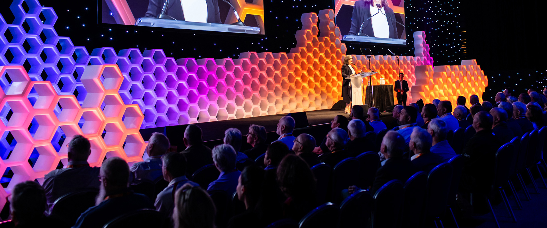 A woman speaks at a podium on a stage with a colorful hexagonal background, addressing a large audience seated in a dimly lit hall.