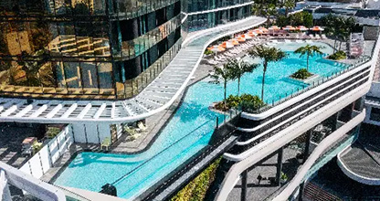 Aerial view of a multi-level outdoor swimming pool with palm trees and lounge chairs, integrated into a modern glass building.