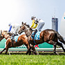 Two jockeys on racehorses galloping side-by-side on a green grass track, with a city skyline in the background.
