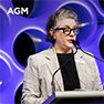A woman with gray hair and glasses speaks at a podium with a microphone, against a purple and blue patterned background with 'AGM' in the corner.