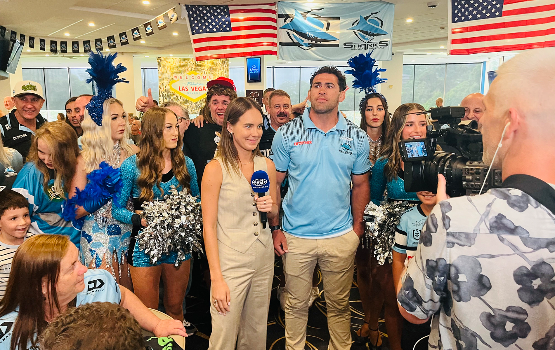 A female reporter interviews a male athlete, surrounded by cheerleaders, fans, and media, with American flags and a Las Vegas sign in the background.