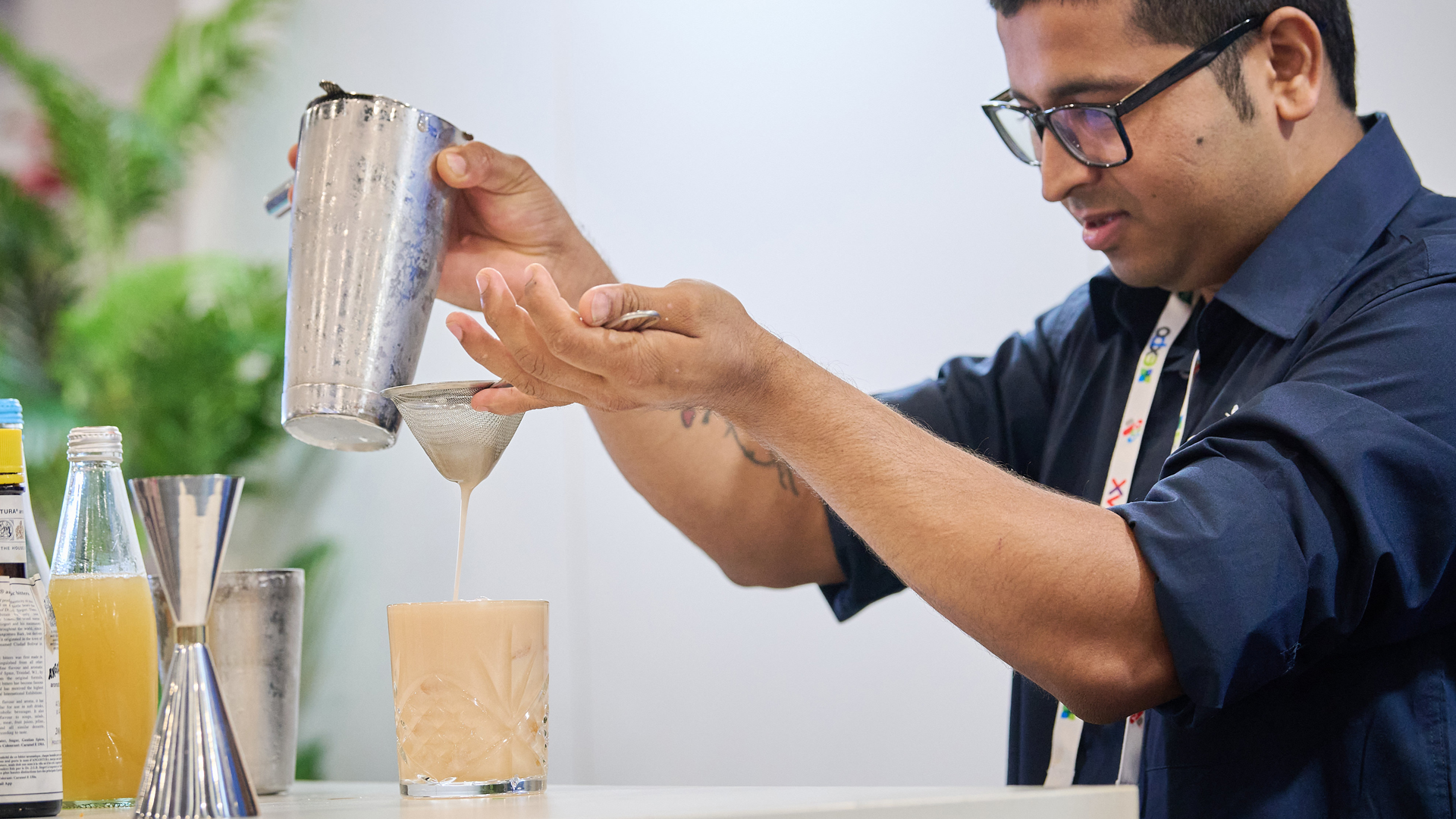 A man in glasses and a dark blue shirt pours a light brown cocktail from a shaker through a strainer into a patterned glass.