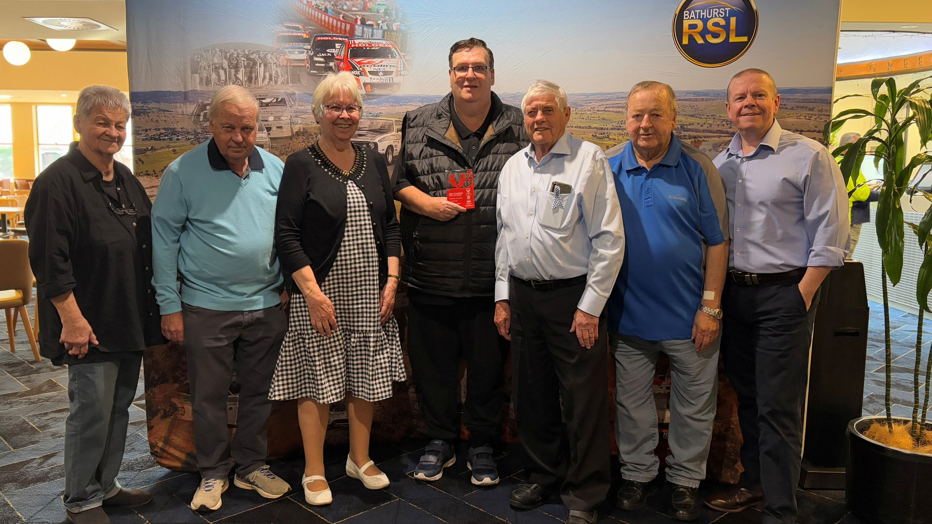 Seven people, five men and two women, stand smiling in front of a racing-themed banner. A man in the center holds a red award plaque for the Bathurst 1000 Hall of Fame.