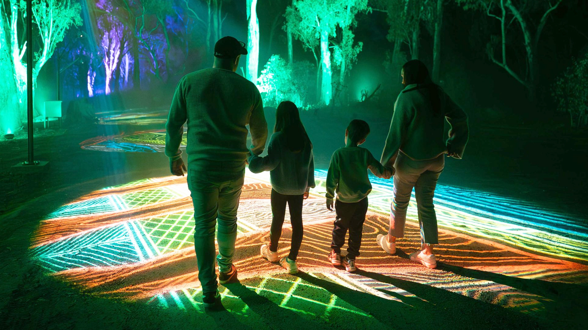 A family of four walks along a path illuminated by colorful light patterns at night, with trees in the background also lit up.