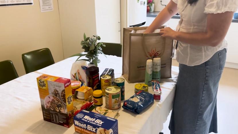 A person in a white shirt and denim skirt is packing groceries and household items into a 'Sydney Community Services' tote bag on a table.