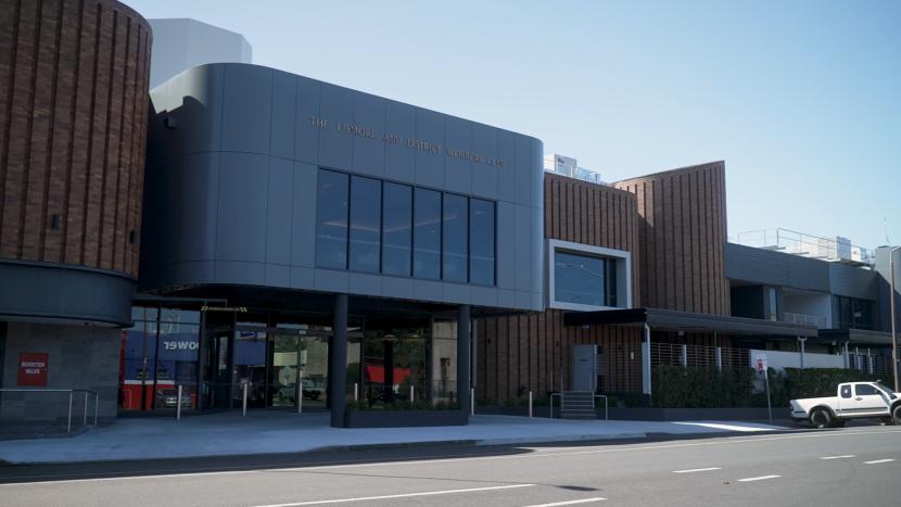 Exterior view of The Lismore and District Workers Club, a modern building with grey curved sections, brown vertical paneling, and large windows, seen from across a street with a parked white pickup truck.