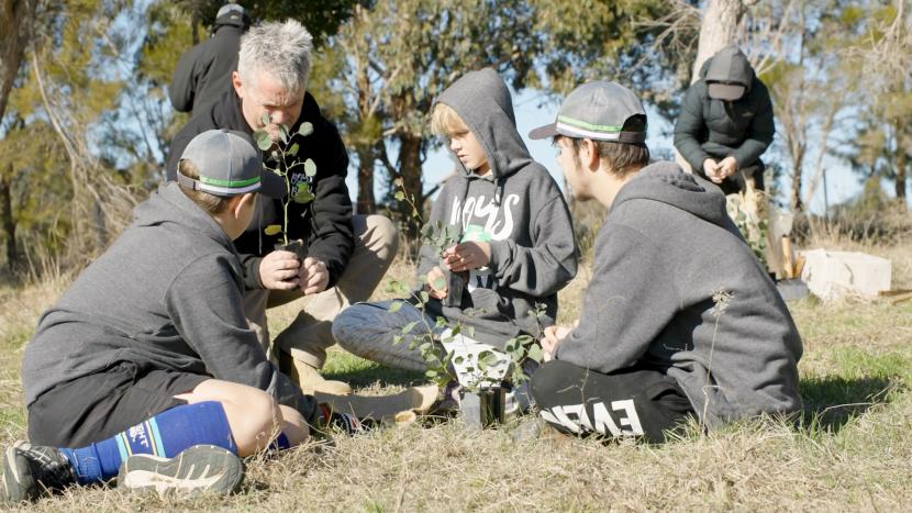 Five people, including adults and children, are planting small trees in a grassy outdoor area.