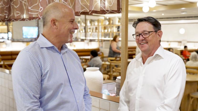 Two smiling men, one in a blue checkered shirt and one in a white shirt, face each other in a restaurant.