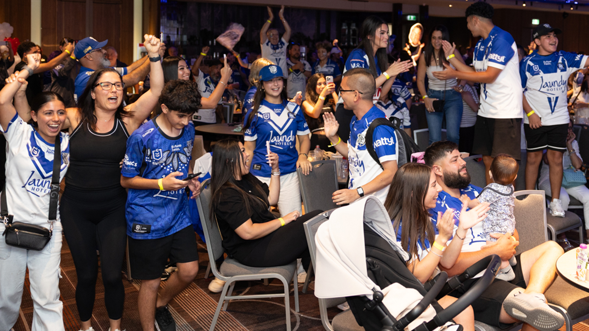A large group of people, many wearing blue and white sports jerseys, cheering and celebrating enthusiastically in an indoor setting.