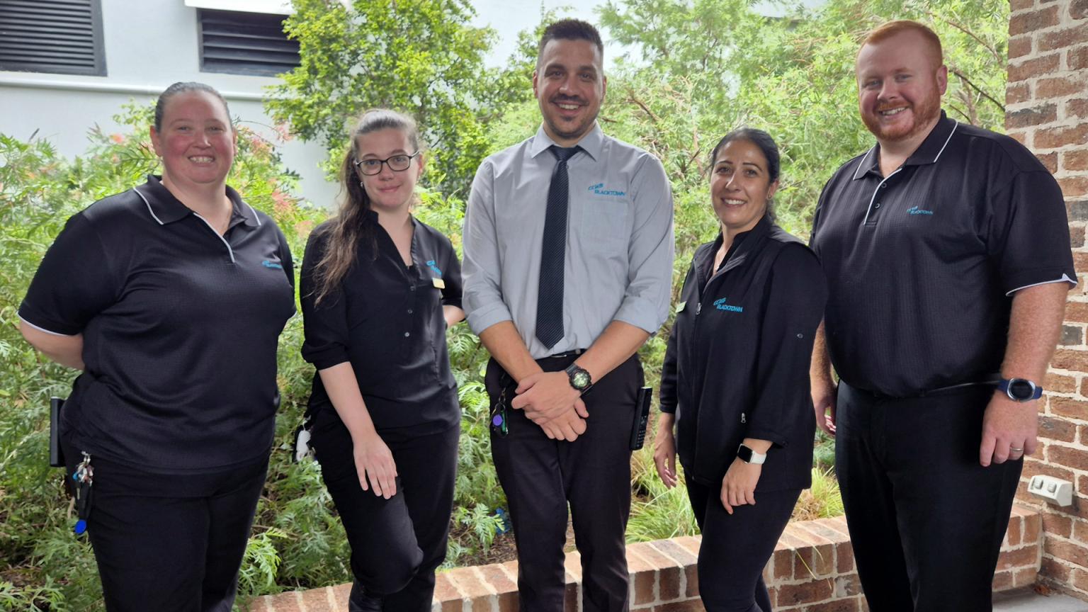 Five people, three women and two men, in branded uniforms, smiling outdoors in front of greenery and a building.