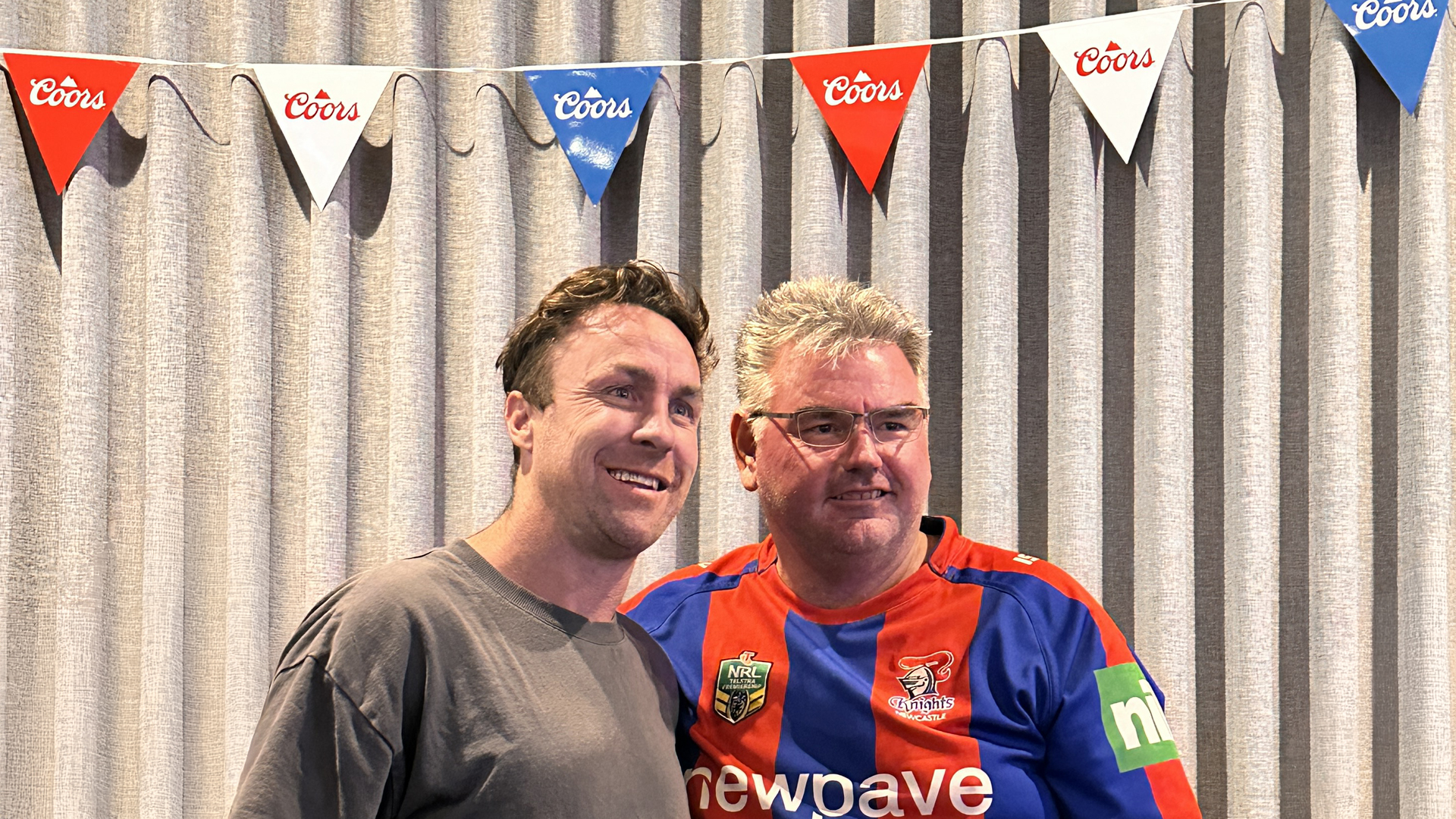 Two smiling men, one in a grey t-shirt and the other in a red and blue sports jersey, stand in front of a striped curtain with Coors bunting above.