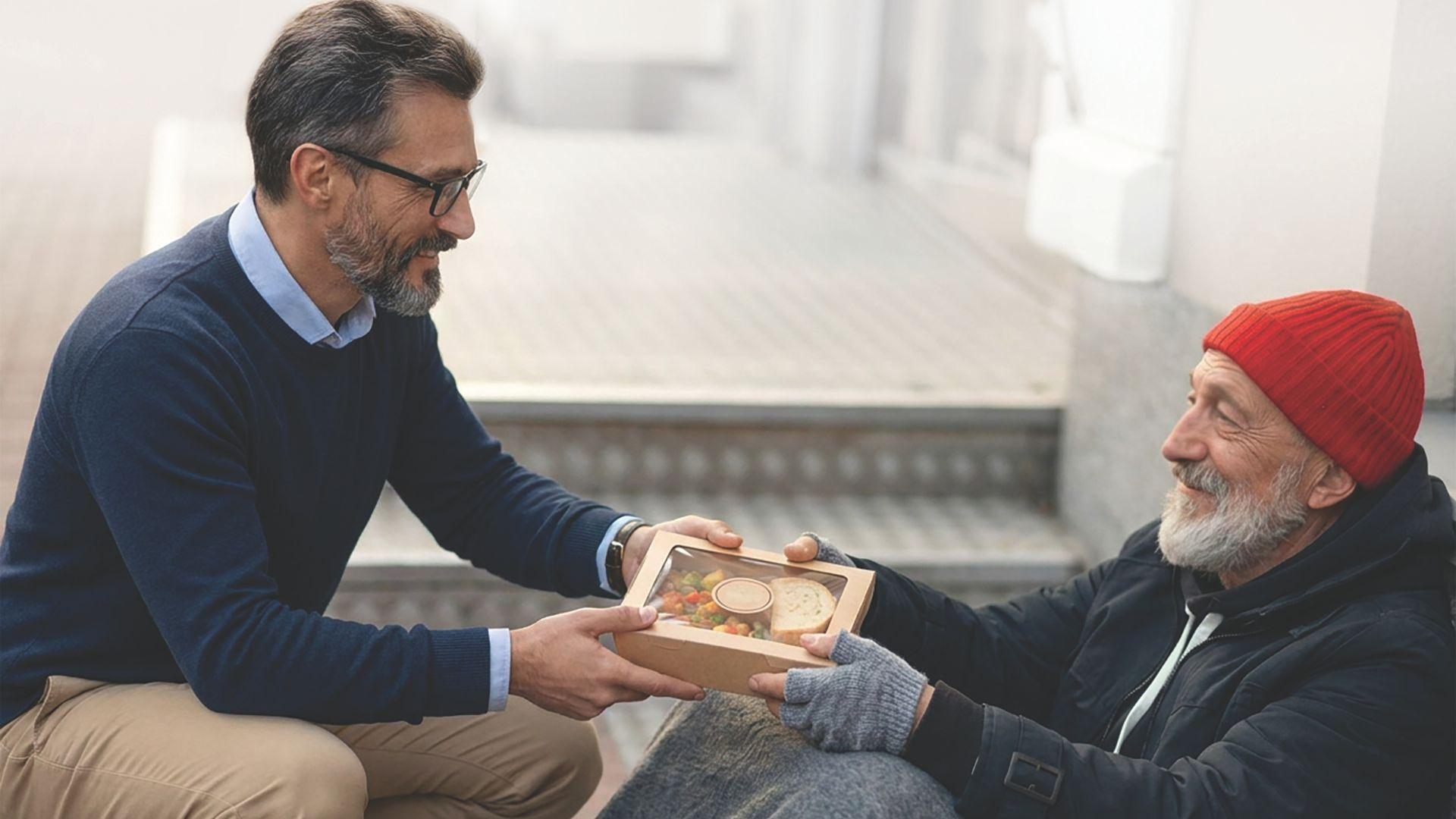 A man in a blue sweater offers a meal in a container to an older man in a red beanie and dark jacket, who is seated on the ground.