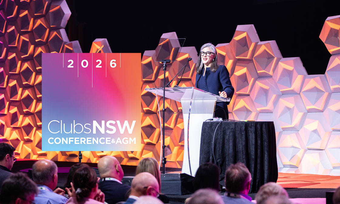 A woman in a dark jacket and glasses speaks at a clear podium on a stage. A large screen behind her displays 'ClubsNSW CONFERENCE & AGM' and '2026'. Geometric wall panels are in the background, and an audience is visible in the foreground.