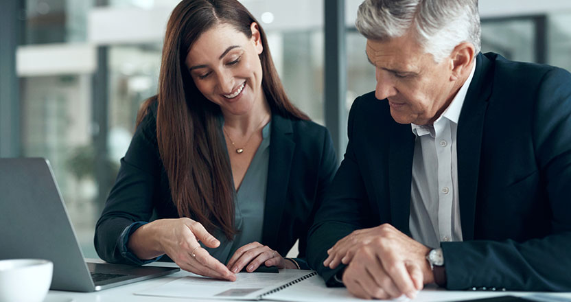 A smiling woman points to a document on a desk while a man in a suit looks on.