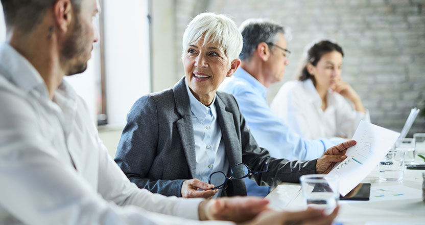 A smiling older woman in a blazer holds documents and glasses, talking to a man during a business meeting with other colleagues in the background.