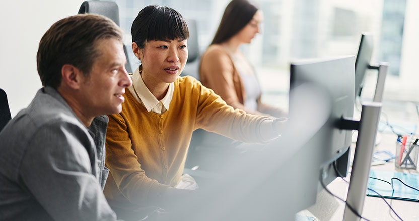 Two colleagues, a man and a woman, collaborate at a computer in an office, with the woman pointing at the screen. Another person works in the blurred background.