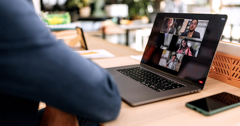 A person's back and arm are visible as they sit at a desk, looking at a laptop screen displaying a video call with six participants. A smartphone and an orange crate are also on the desk.