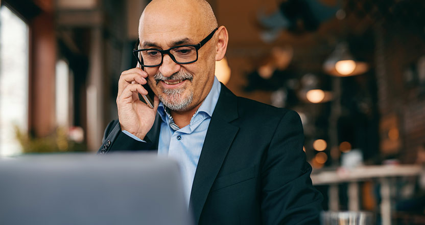 A smiling bald man in glasses talks on a phone while looking at a laptop in a blurred indoor setting.