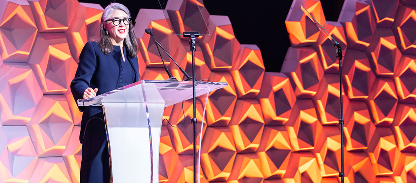 Woman with gray hair and glasses speaking at a clear podium with two microphones, against a colorful geometric background.