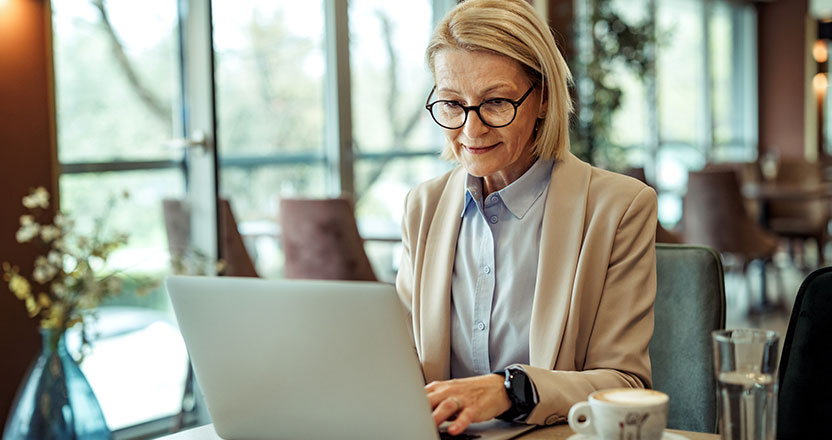 A blonde woman in glasses and a beige blazer types on a silver laptop at a cafe table with coffee and water.