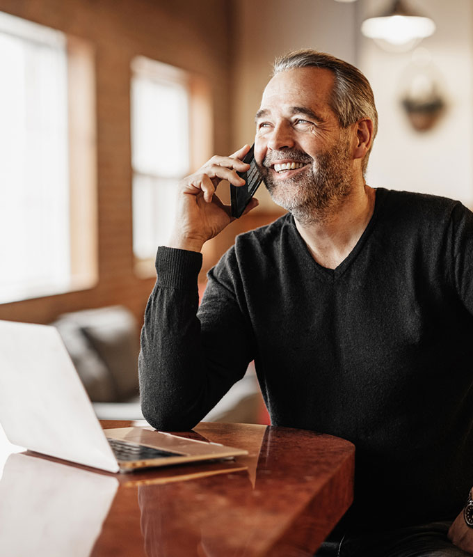 A smiling man with graying hair on a phone call, with a laptop open on a wooden table.