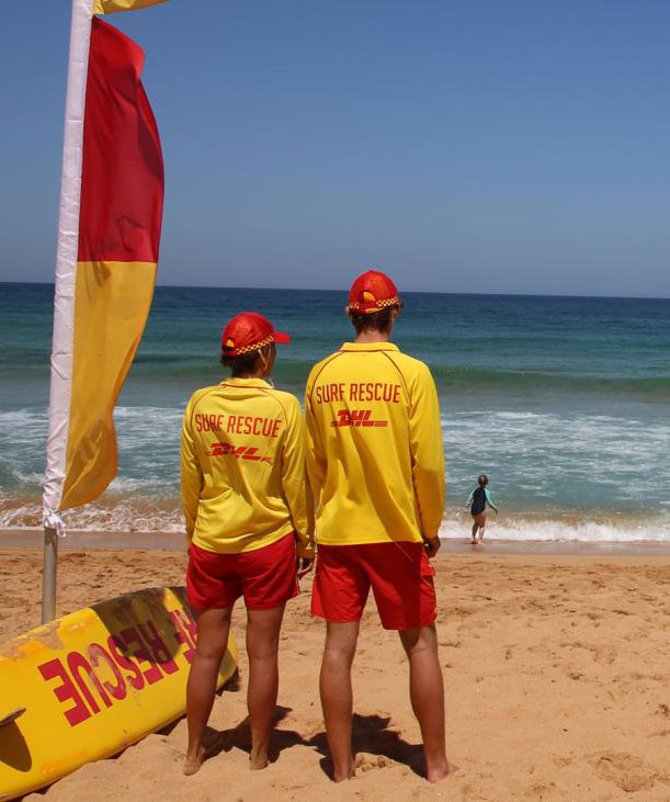 Two surf rescuers in yellow shirts and red shorts stand on a sandy beach, looking out at the blue ocean. A child is in the water in the distance.