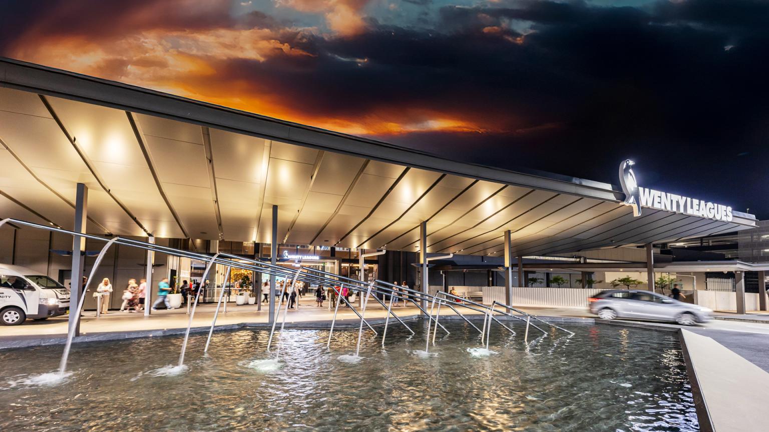 Modern building entrance of Wenty Leagues at night, with a water feature, illuminated canopy, and a dramatic orange and dark sky.