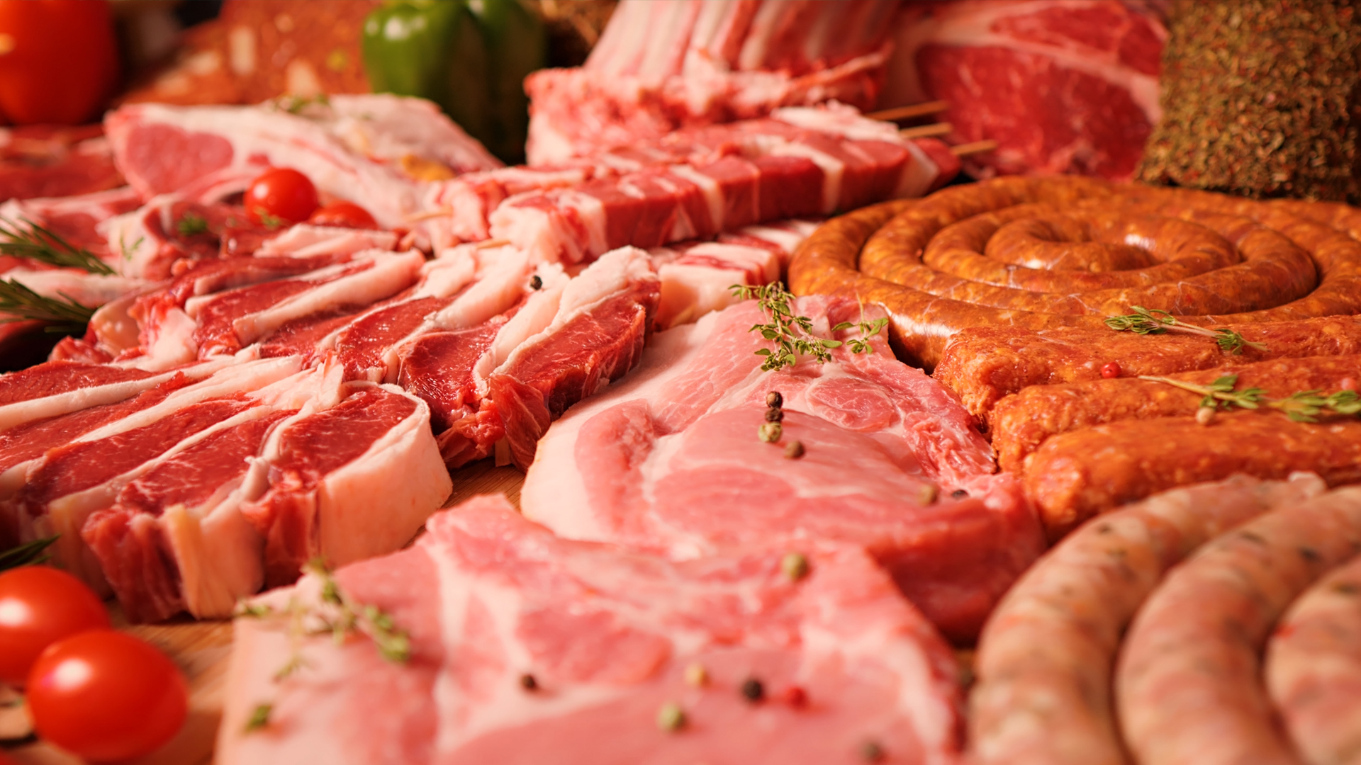 A close-up of a diverse assortment of raw meats, including red meat chops, pink pork steaks, and various sausages, garnished with herbs, peppercorns, and cherry tomatoes.