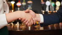 Two people shaking hands in front of a blurred bar counter with beer taps.