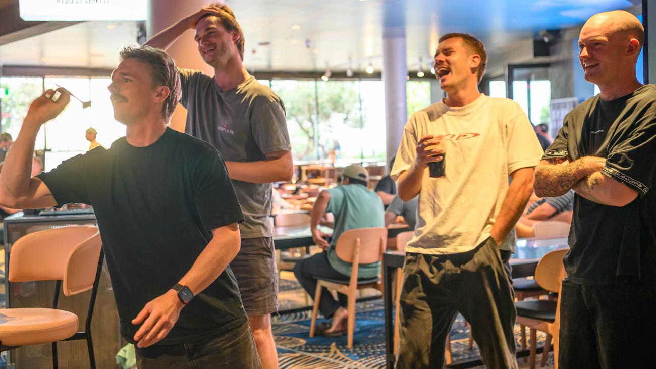 Four men in a casual indoor setting. One man holds a dart, while the others laugh and smile, some holding drinks.