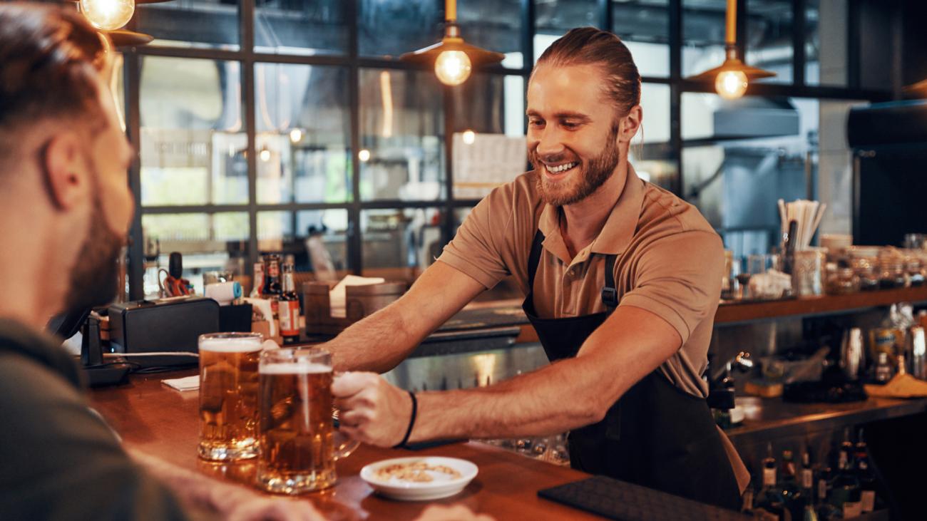 A smiling male bartender serves two mugs of beer to a customer at a wooden bar counter.