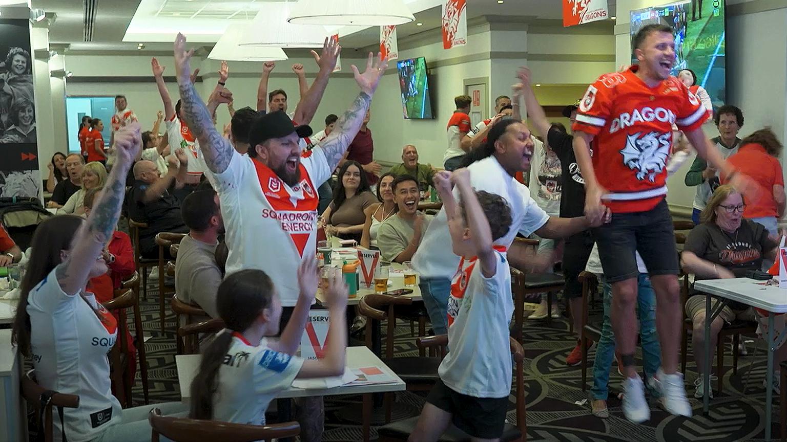 A large group of enthusiastic sports fans in red and white jerseys cheering and jumping indoors, watching a game on a TV screen.