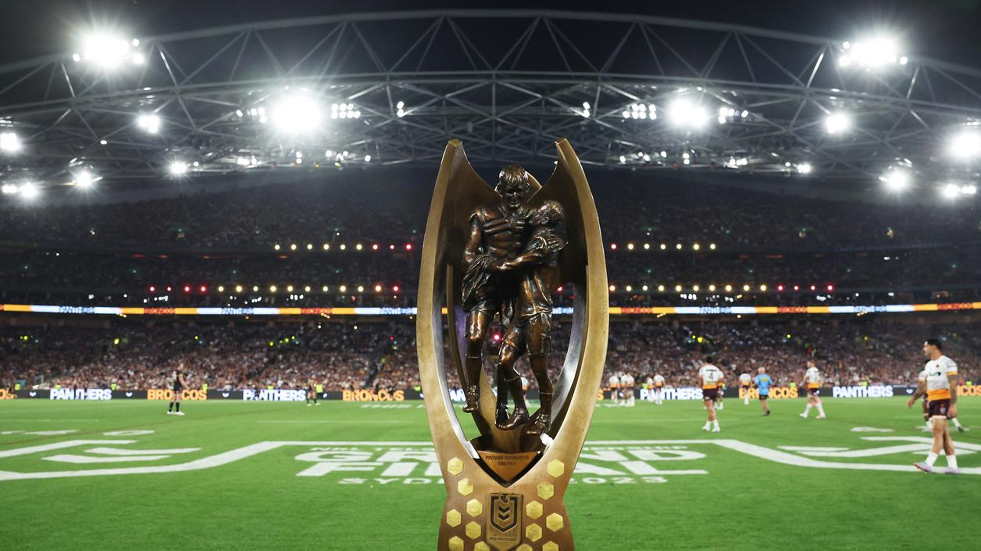 A bronze rugby league trophy with two players on a green field, with a brightly lit stadium and crowd in the background at night.