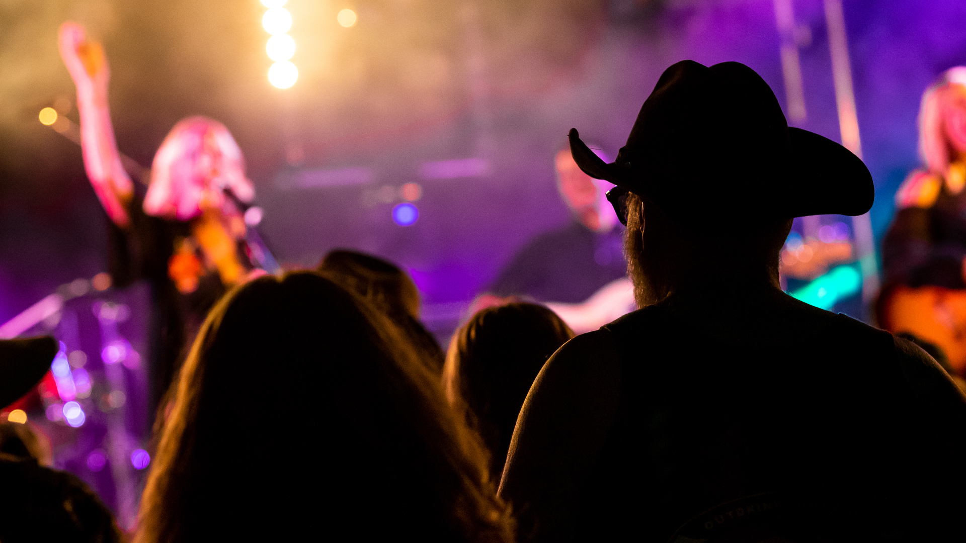 Rear view of a person in a cowboy hat and other audience members at a concert, silhouetted against a brightly lit stage with blurred performers and colorful lights.