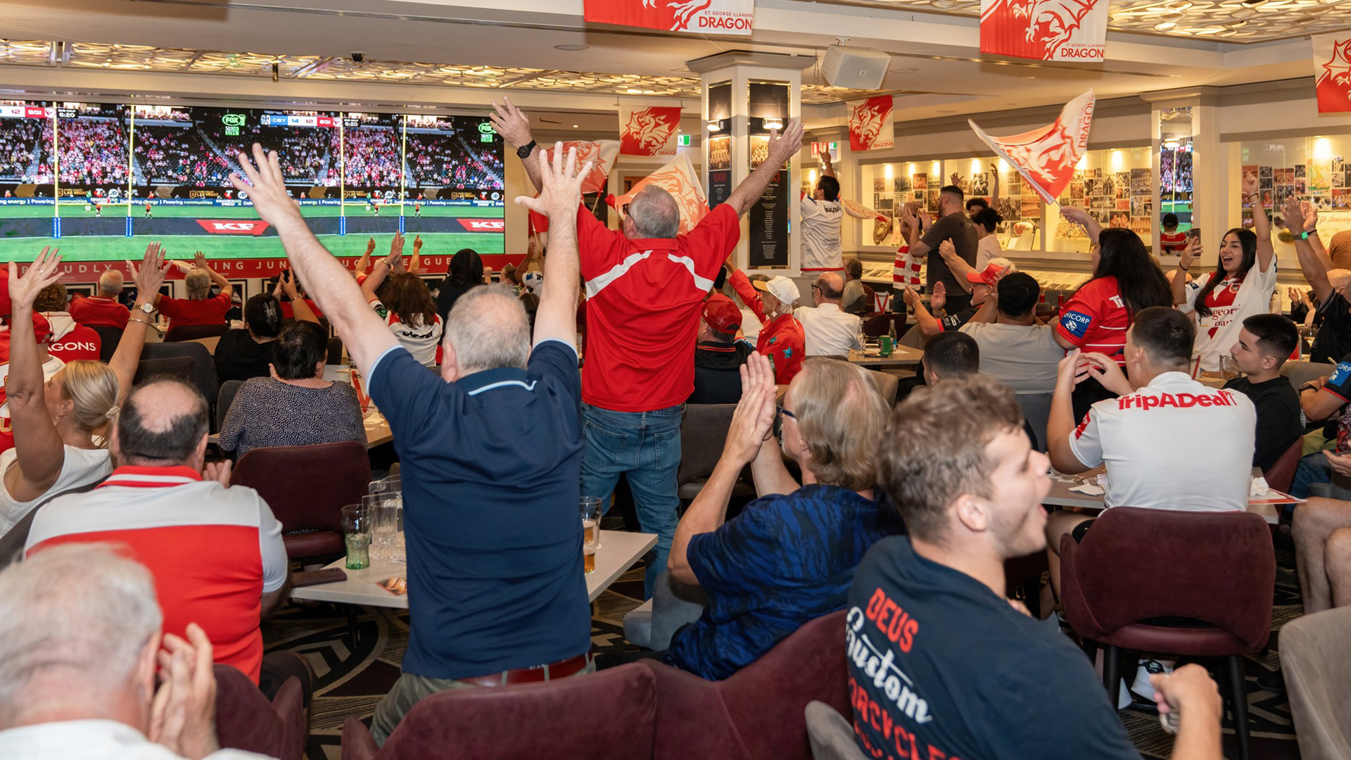 A large group of excited sports fans cheer with raised arms while watching a rugby game on a big screen in a decorated room.
