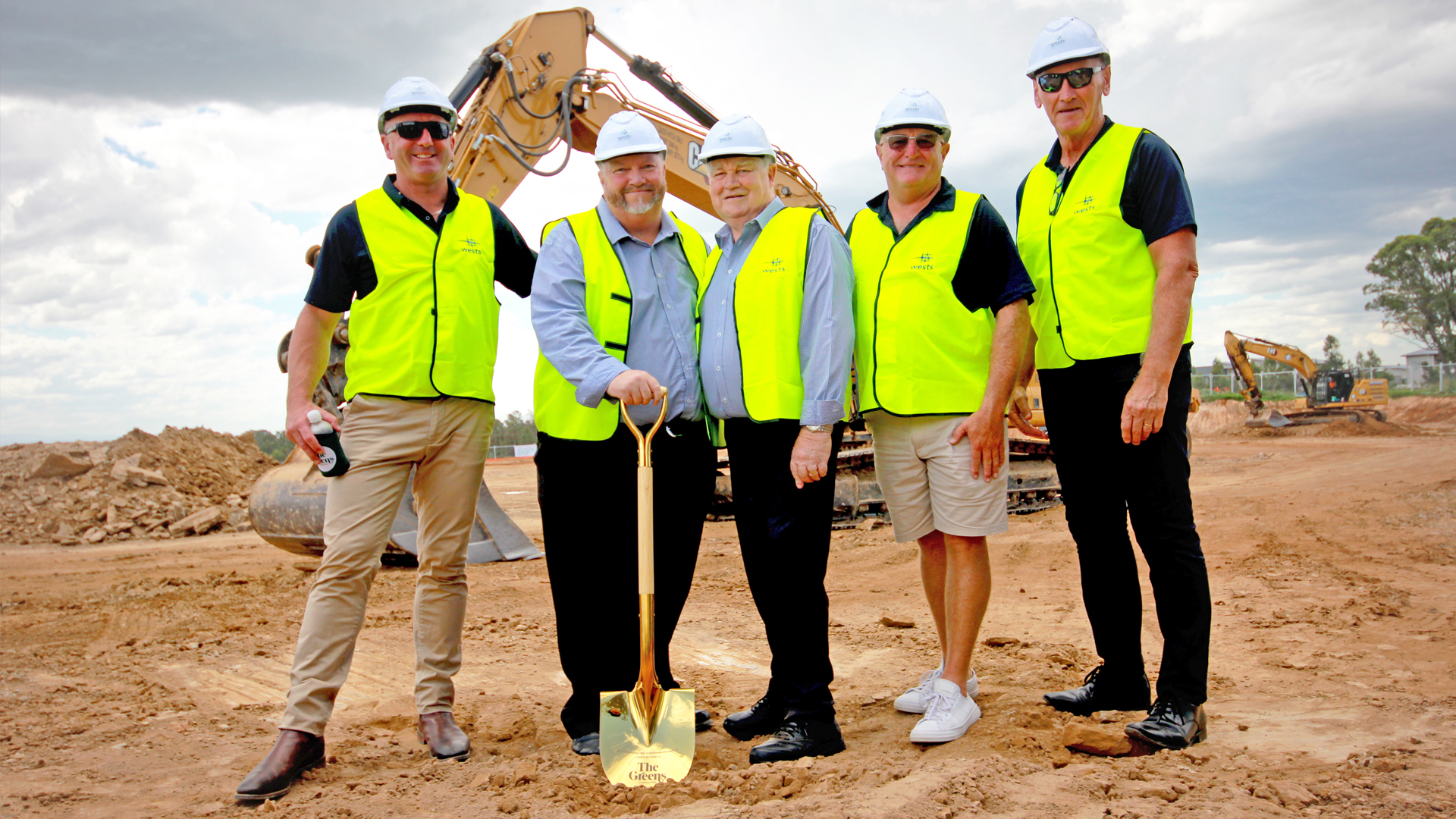 Five men in hard hats and yellow safety vests stand on a dirt construction site with a golden shovel, excavators in the background.