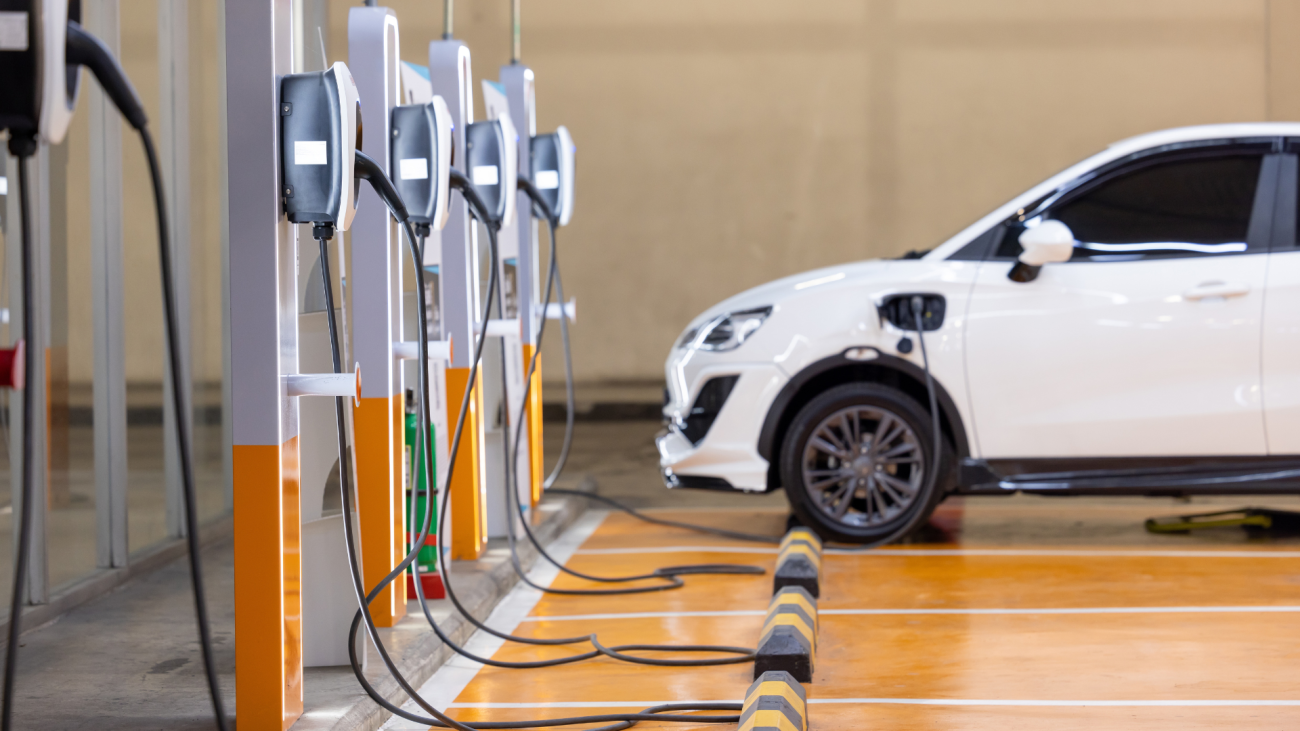 A white electric vehicle is charging at a row of charging stations with orange flooring and parking stops.