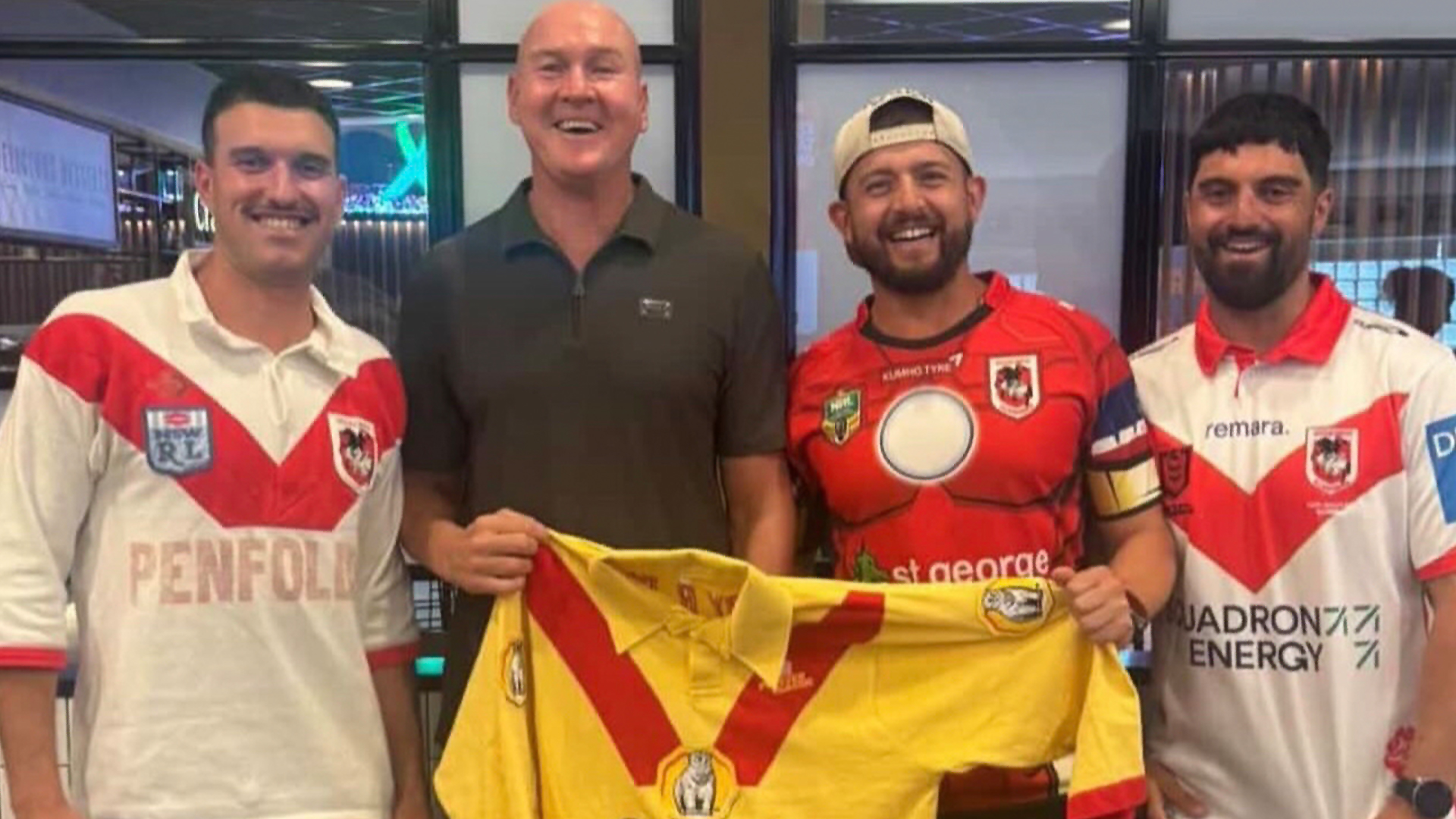 Four smiling men, two in white and red jerseys, one in a red Iron Man shirt, and one in a dark polo, holding up a yellow and red rugby jersey.