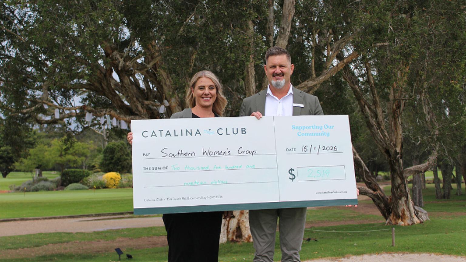 A woman and a man holding a large novelty check from Catalina Club for $2,519 made out to Southern Women's Group, standing on a green lawn with trees in the background.