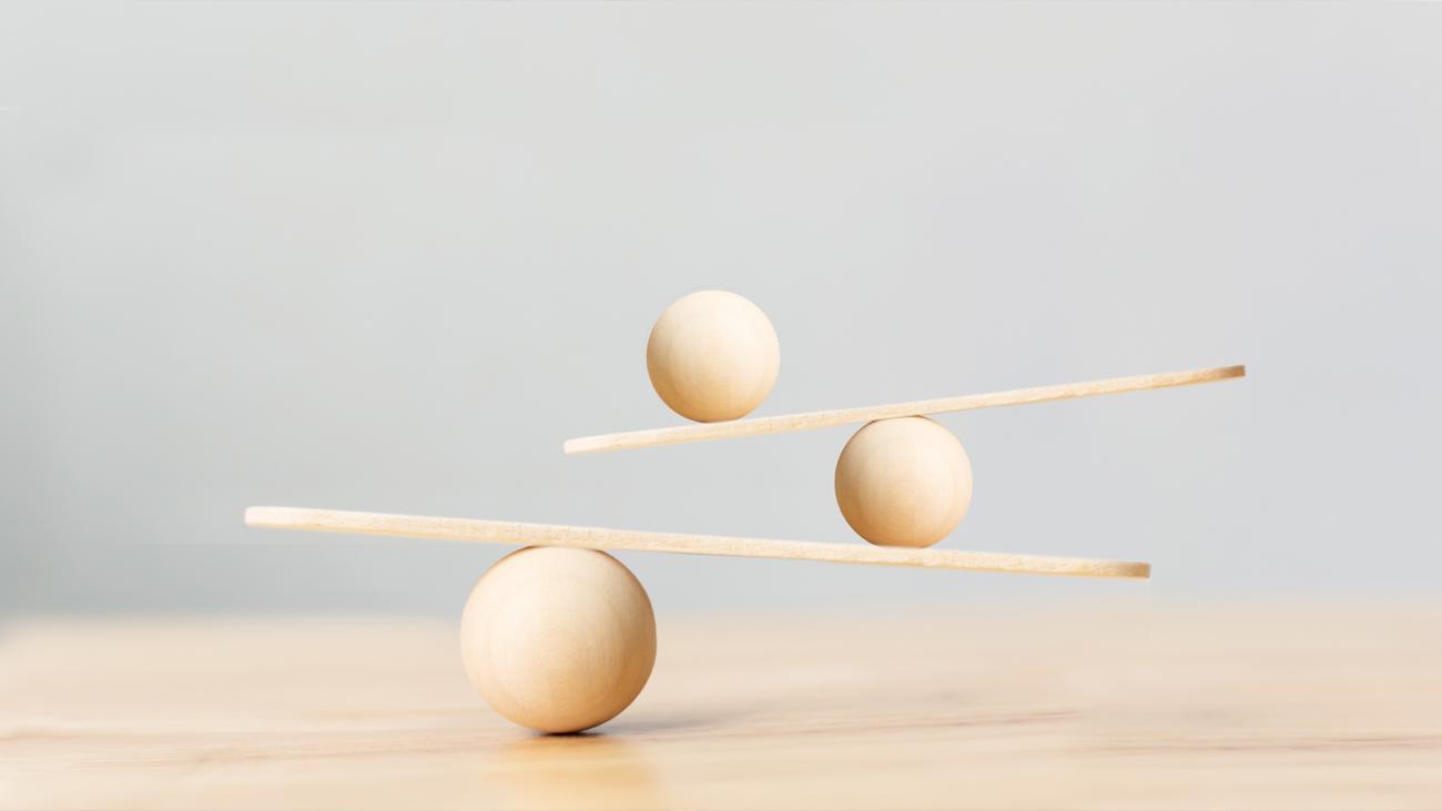 Three light wooden spheres and two wooden sticks arranged in a balancing act on a light wooden surface, against a light gray background.