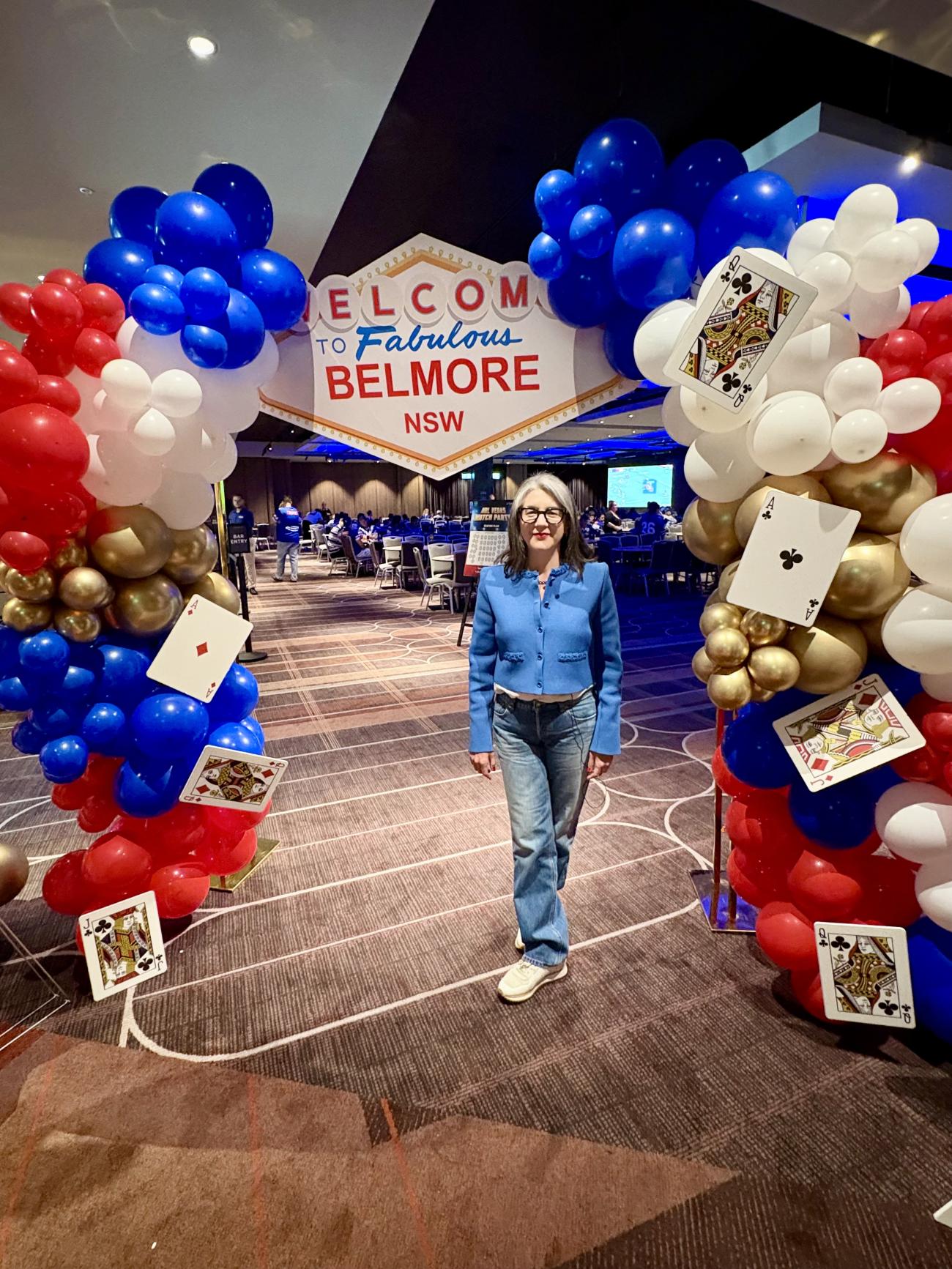 A woman stands smiling under a balloon archway decorated with playing cards, with a 'Welcome to Fabulous Belmore NSW' sign above her, leading into an event hall.