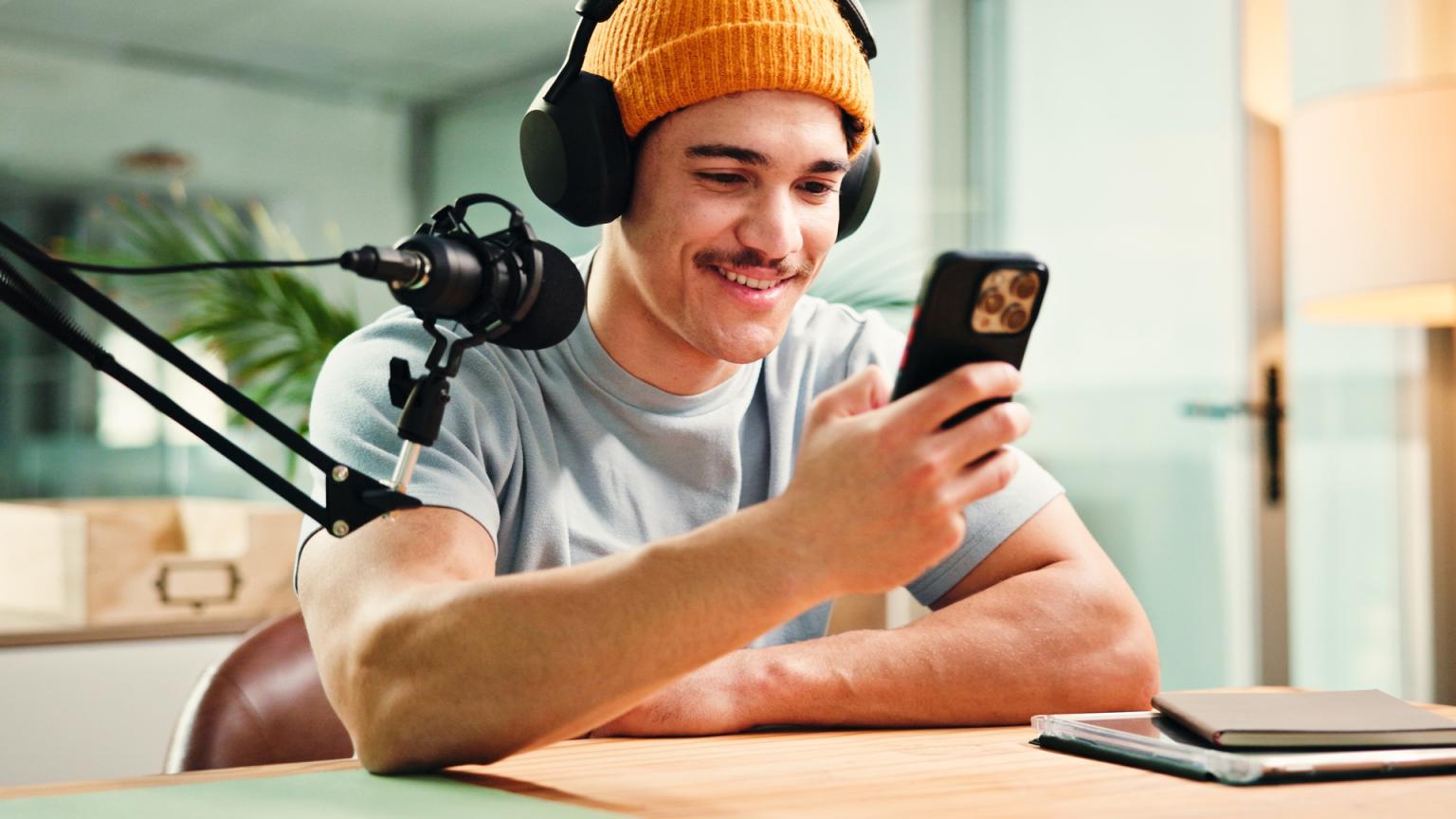 A smiling young man in an orange beanie and headphones looks at his smartphone, with a microphone and recording setup nearby.
