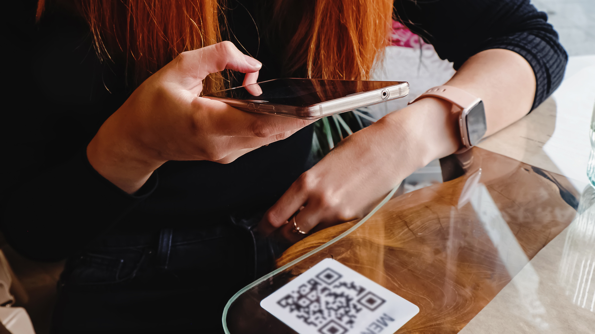 A person holds a smartphone over a QR code on a glass-topped table, with a smartwatch on their wrist.